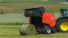 Presse à balles rondes KUHN FB 3130 en train de produire une balle d'ensilage liée par filet dans une région vallonnée.